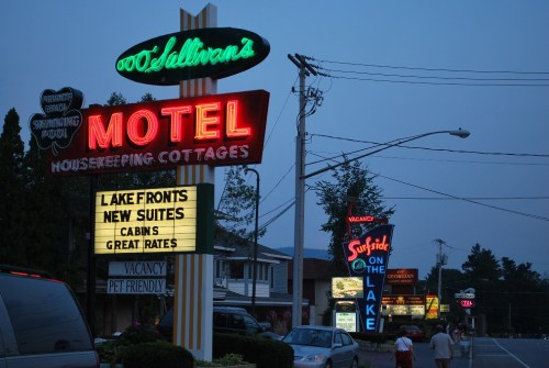 Neon signs for motels and cabins along a road, with trees and a lake in the background during twilight.