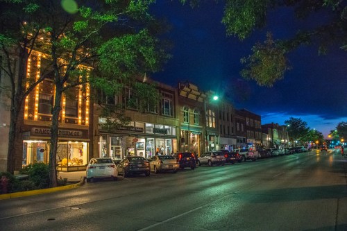 A charming street scene at dusk, featuring shops with lights and parked cars under a blue sky.