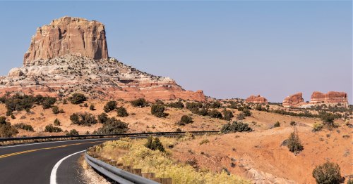 A winding road leads through a desert landscape with a prominent rock formation and distant red cliffs under a clear sky.