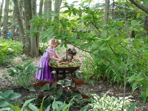 A child in a purple dress tends to a flower garden surrounded by lush greenery and trees.