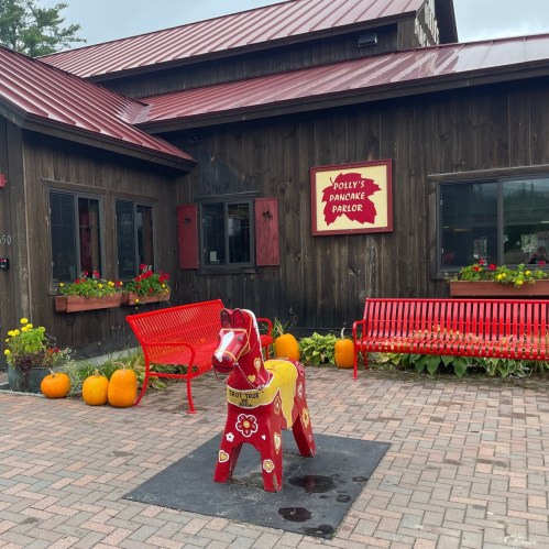 A red wooden horse statue stands outside Polly's Pancake Parlor, surrounded by flowers and pumpkins on a brick path.
