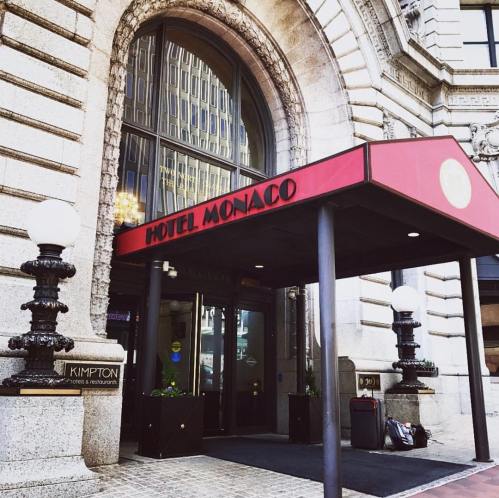 Entrance of Hotel Monaco with a red awning and decorative columns, featuring large windows and a sign.