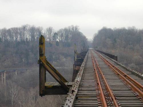 A long, abandoned railway bridge stretches into the distance, surrounded by trees and a foggy landscape.