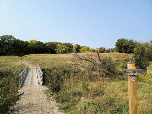 A wooden bridge leads to a grassy hill with trees under a clear blue sky. A trail sign is visible in the foreground.
