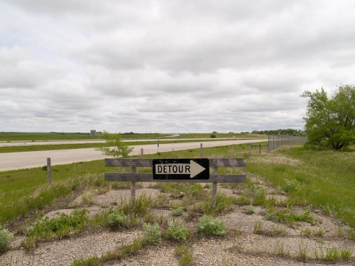 A weathered detour sign points right, with a grassy roadside and a cloudy sky in the background.
