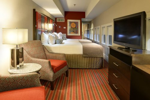 A hotel room featuring two beds, a chair, a TV, and colorful striped carpet. Bright and modern decor.