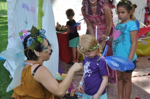 A woman decorates a young girl with fairy wings, while two other children watch in a colorful outdoor setting.