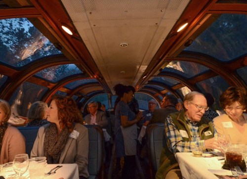 A dining car interior with guests enjoying a meal, soft lighting, and a view of the outside through large windows.