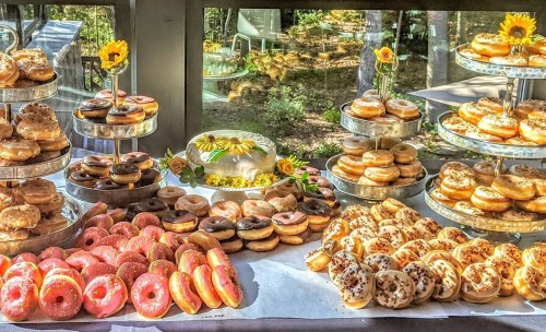 A colorful display of assorted donuts on tiered stands, with a cake and sunflowers in a bright, natural setting.