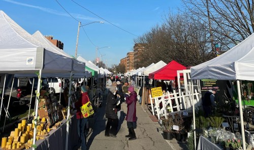 A bustling outdoor market with white tents, vendors, and shoppers under a clear blue sky.