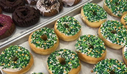 A tray of colorful donuts topped with green sprinkles and icing, alongside chocolate and pink frosted donuts.