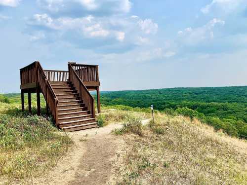 A wooden observation deck on a grassy hill, overlooking a lush green landscape under a partly cloudy sky.