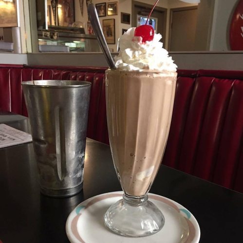 A tall glass of chocolate milkshake topped with whipped cream and a cherry, next to a metal cup on a diner table.