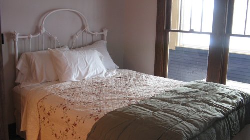 A cozy bedroom featuring a white metal bed with a quilted cover and large windows letting in natural light.