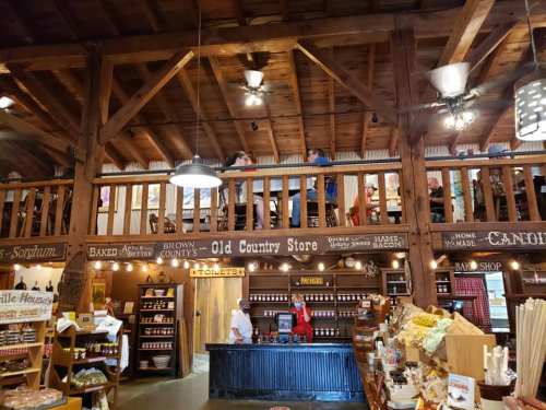 Interior of a rustic country store with wooden beams, shelves of goods, and customers on a balcony above.