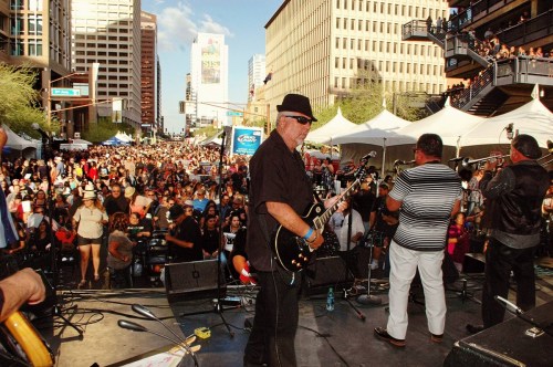A lively outdoor concert scene with a large crowd, musicians on stage, and city buildings in the background.