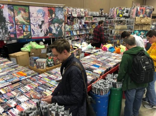 A busy convention scene with attendees browsing a large display of anime merchandise and collectibles.