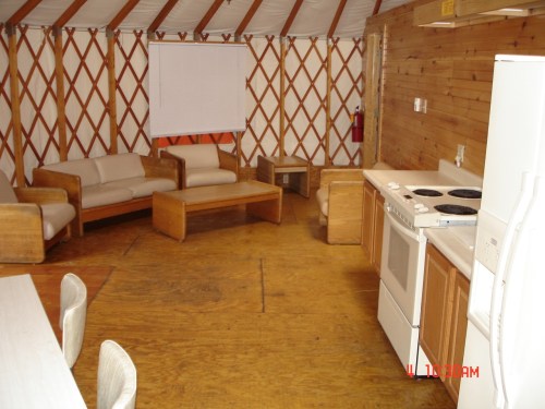 Interior of a yurt featuring wooden furniture, a kitchen area, and a cozy seating arrangement.