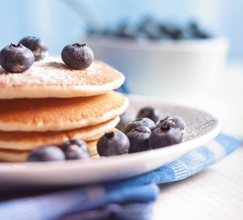 A stack of pancakes topped with blueberries, served on a plate with more blueberries in the background.