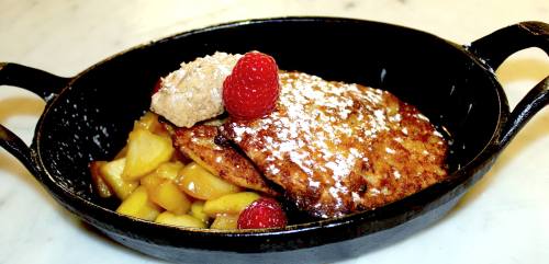 A cast iron dish with two pancakes topped with powdered sugar, served with sautéed apples and a dollop of cream.