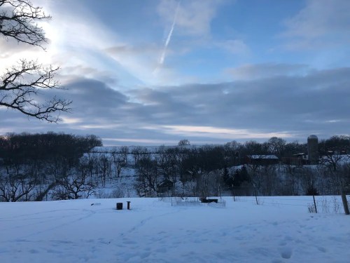 A snowy landscape with trees, a barn, and a silo under a cloudy sky, creating a serene winter scene.
