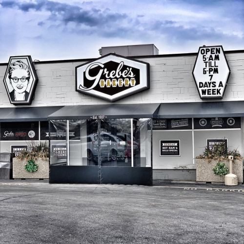 Exterior of Grebe's Bakery, featuring a sign with hours and a vintage-style logo, set against a cloudy sky.