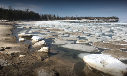 A frozen shoreline with ice patches scattered across a calm lake, surrounded by trees and a clear blue sky.