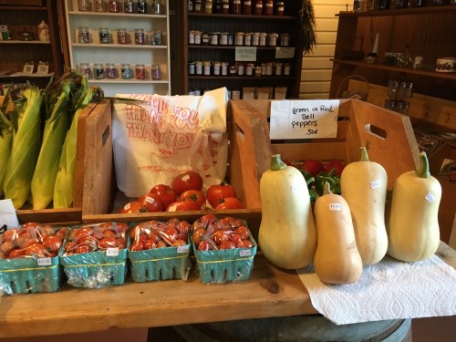 A wooden display of fresh produce, including corn, tomatoes, bell peppers, and butternut squash in a market setting.