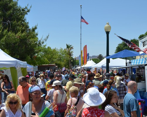 A bustling outdoor market with crowds of people, colorful tents, and flags under a clear blue sky.
