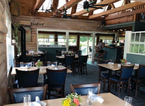 Cozy restaurant interior with wooden beams, tables set for dining, and a bar in the background. Bright and inviting atmosphere.