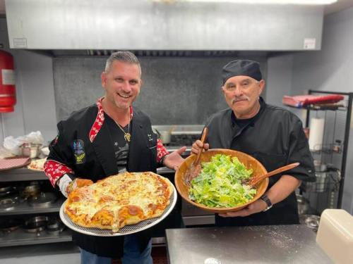 Two chefs in a kitchen, one holding a large pizza and the other a bowl of salad, both smiling at the camera.