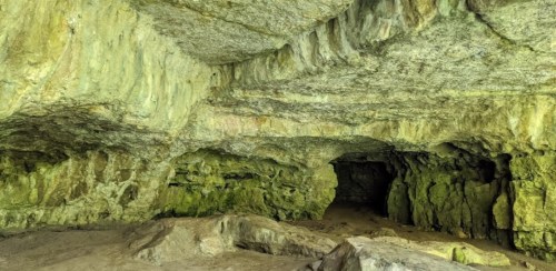 A spacious cave interior with textured rock walls and a sandy floor, illuminated by soft, natural light.