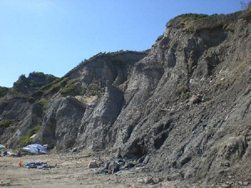 Rocky cliffside with steep slopes, partially covered in vegetation, overlooking a sandy beach. Clear blue sky above.