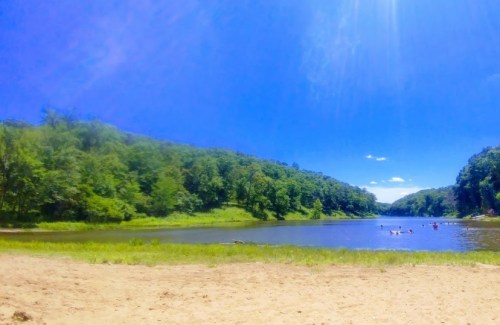 A serene river scene with lush green hills, a sandy shore, and people enjoying the water under a bright blue sky.