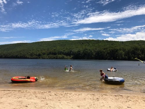 A sunny beach scene with people swimming and playing in the water, surrounded by green hills and a clear blue sky.