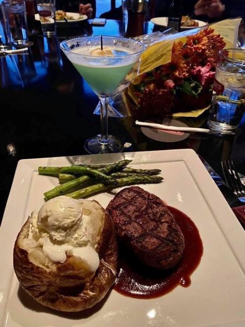 A plate featuring a grilled steak with sauce, a baked potato topped with sour cream, asparagus, and a blue cocktail.