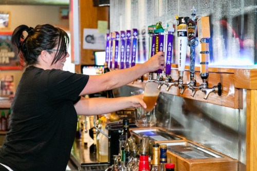 A bartender pours a beer from a tap at a bar, with various beer taps and bottles visible in the background.
