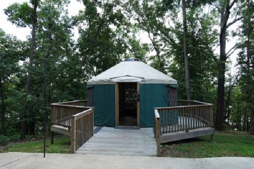 A wooden yurt with a green exterior, surrounded by trees, featuring a deck and steps leading to the entrance.