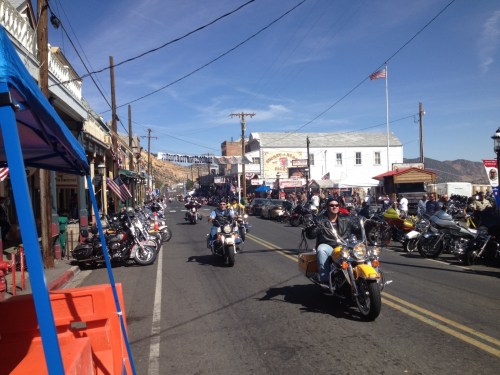 A busy street scene with motorcycles riding through a historic town, lined with buildings and flags under a clear blue sky.