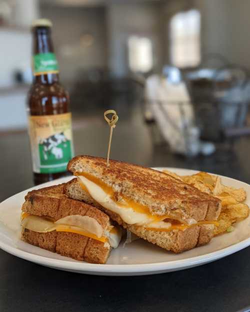 A grilled cheese sandwich with melted cheese, pickles, and a side of chips, accompanied by a bottle of soda.