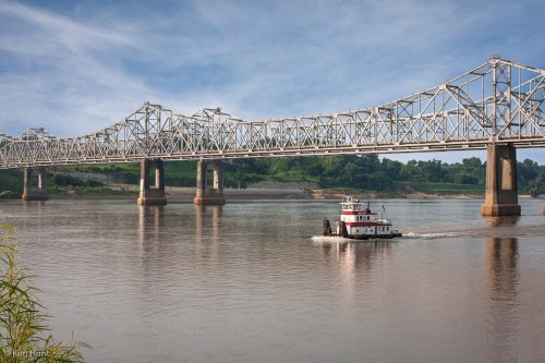 A tugboat navigates the river beneath a large steel bridge on a clear day.