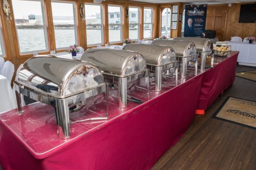A buffet setup with silver chafing dishes on a red tablecloth, overlooking a waterfront view through large windows.