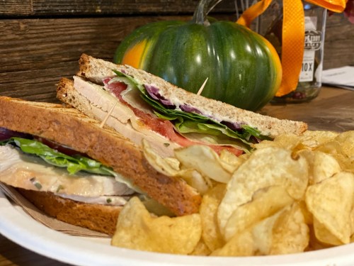 A sandwich with turkey, lettuce, tomato, and purple cabbage, served with potato chips and a decorative pumpkin in the background.