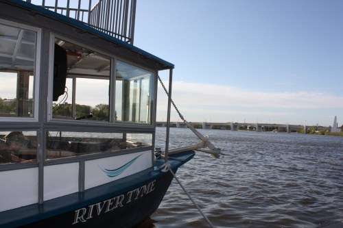 A boat named "RIVER TYE" is moored on a calm river, with a bridge visible in the background under a clear sky.