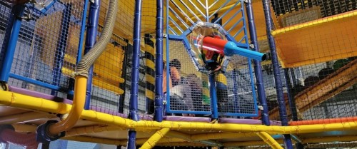 Colorful indoor playground structure with climbing areas and slides, children playing inside a mesh enclosure.