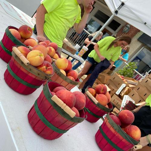 Baskets of peaches on a table at a market, with people in bright shirts and a building in the background.