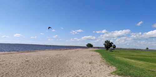 A sandy beach by a calm lake under a blue sky with scattered clouds and green grass in the foreground.