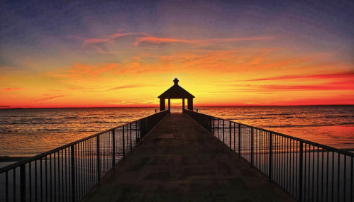 A serene sunset over the ocean, viewed from a wooden pier leading to a small gazebo. Vibrant colors fill the sky.
