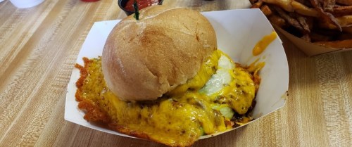 A cheesy burger with a toasted bun, served with a side of fries and dipping sauce on a wooden table.