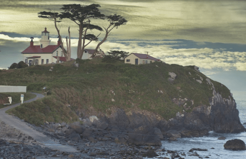A lighthouse on a rocky cliff, surrounded by trees, with a cloudy sky and ocean in the background.